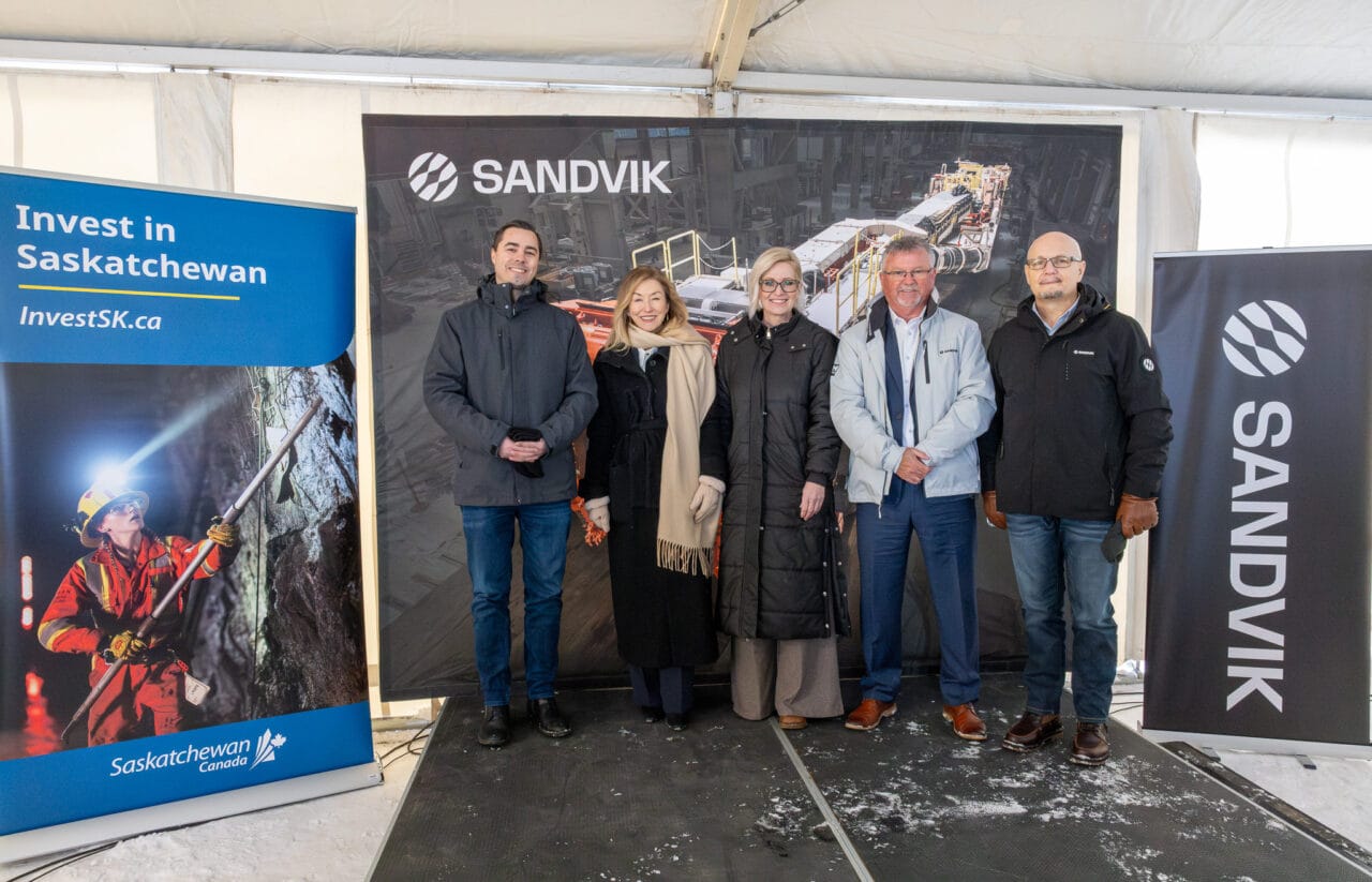 Government and company officials stand together inside a white event tent at the Sandvik groundbreaking ceremony in Saskatchewan, gathered in front of event signage as they mark the announcement of the new mining equipment facility.
