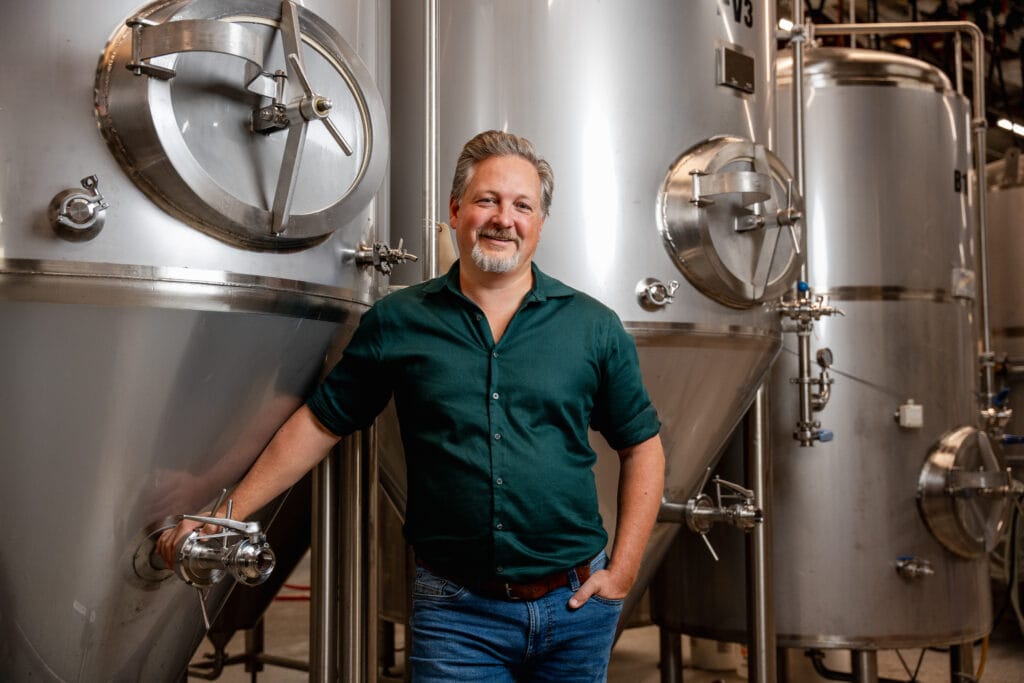 Shawn Moen standing in front of large stainless steel barrels used for brewing beer in a brewery.