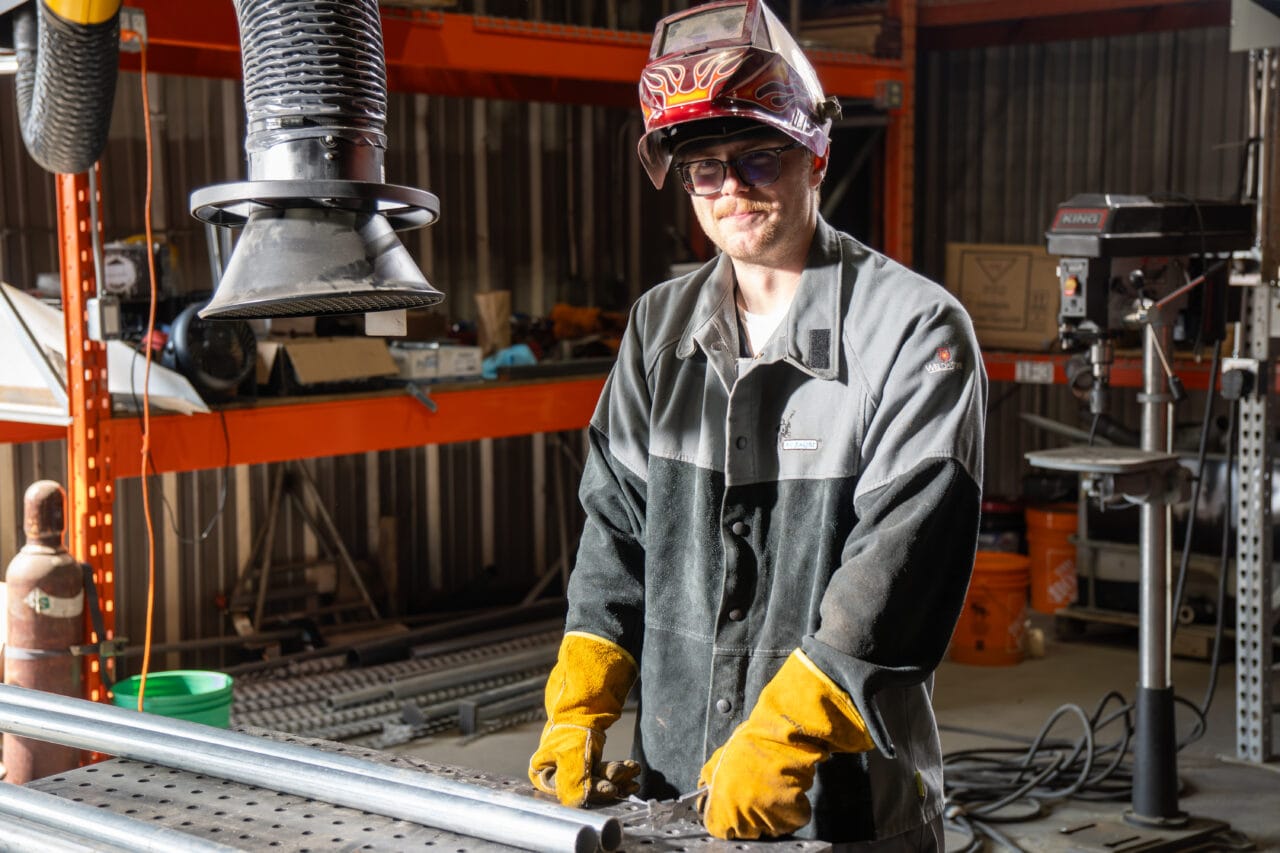 Welder working at a shop bench in an industrial setting, focused on metal fabrication work with tools and equipment visible in a workshop environment.