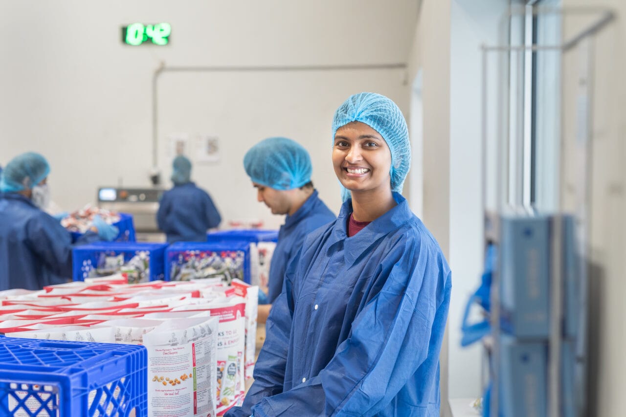 Worker in a food processing facility packaging products on a production line. The employee is wearing appropriate safety and sanitation equipment while preparing goods for distribution in a clean manufacturing environment.
